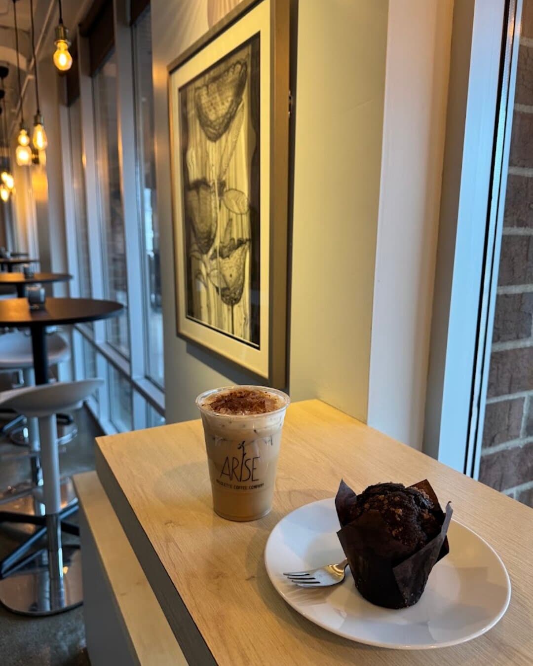 An iced latte and a chocolate muffin on the window bar, framed art on the wall.